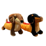 Two assorted dachshund plush toys standing on a wooden table. One plush features brown and cream colors, while the other has a black and tan design. Both have long bodies, floppy ears, embroidered smiles, and soft plush fabric.