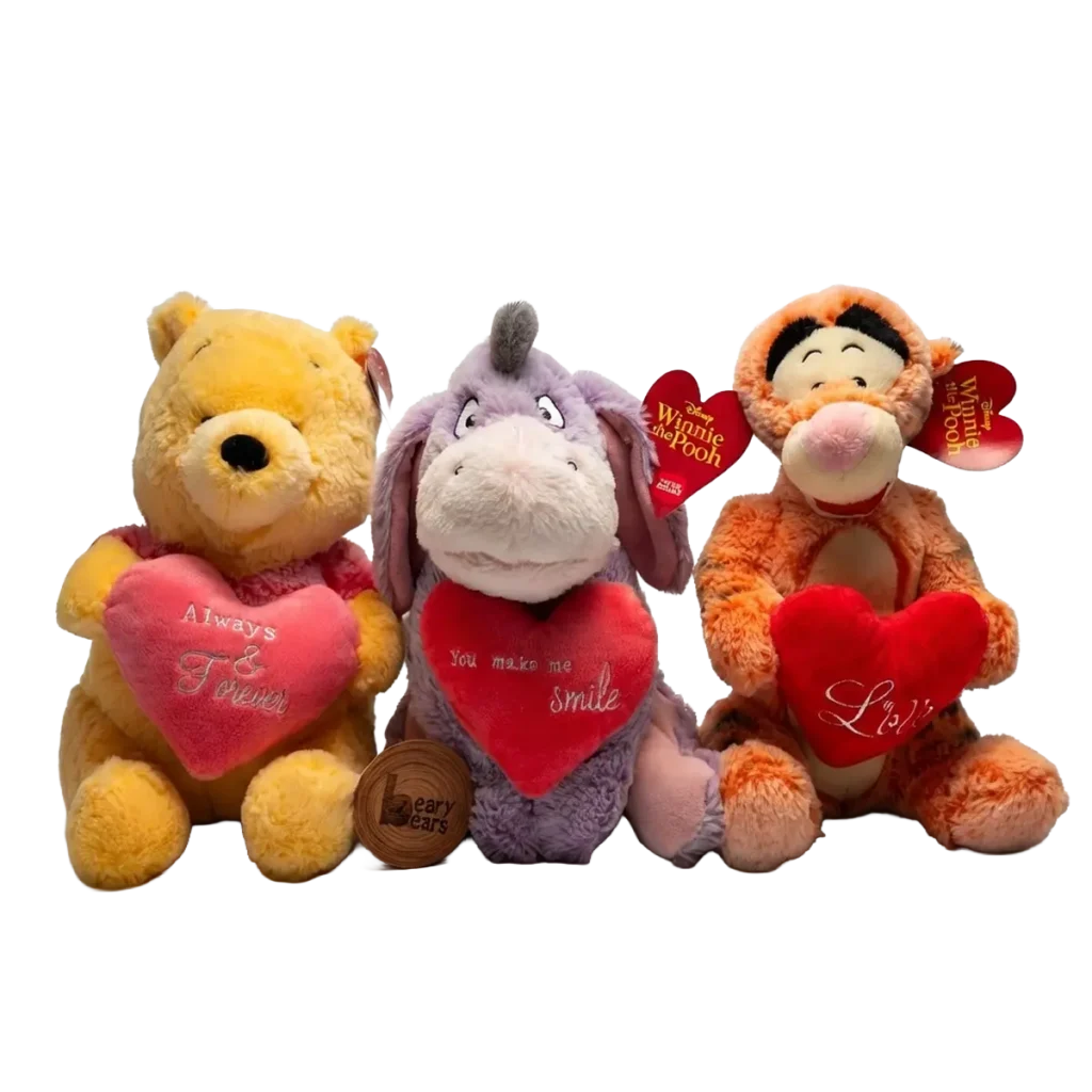 Three plush toys sitting together holding red heart cushions: a yellow bear, a purple donkey, and an orange tiger plush.
