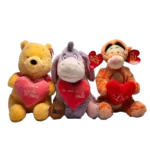 Three plush toys sitting together holding red heart cushions: a yellow bear, a purple donkey, and an orange tiger plush.